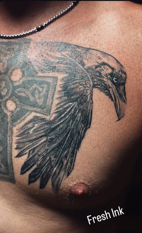 Tattoo eines Raben in dunkler Tinte auf der Haut, mit keltischem Kreuz im Hintergrund.