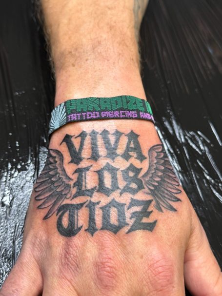 Tattoo auf einer Hand mit dem Schriftzug „VIVA LOS TIOZ“ und einem bunten Armband.