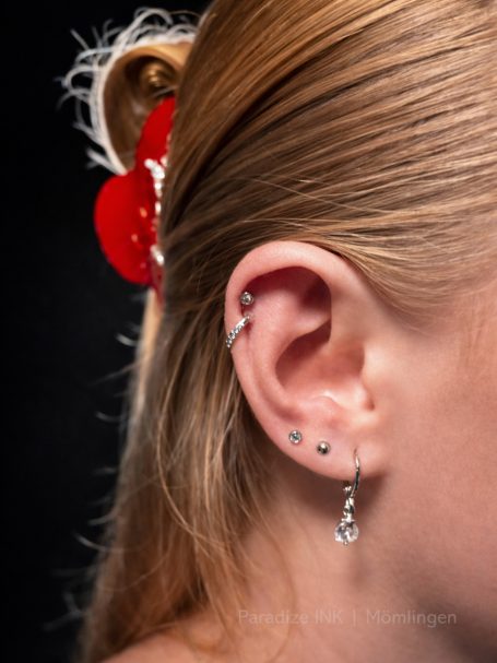 Detailaufnahme eines Ohrs mit mehreren Ohrringen und einer roten Haarnadel im Haar.