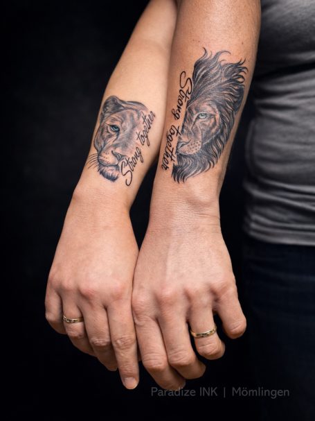 Zwei Arme mit Löwen- und Tiger-Tätowierungen, nahe beieinander gehalten.