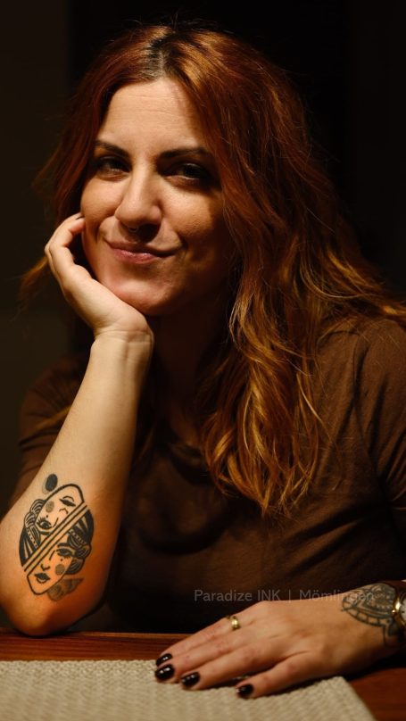 Lächelnde Frau mit langen, rotbraunen Haaren und einem Tattoo auf dem Arm.