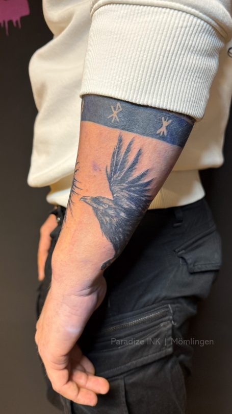 Tattoo eines Vogels, der am Unterarm auf schwarzem Hintergrund abgebildet ist.