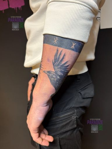 Tattoo eines Krähenkopfes auf dem Unterarm, umgeben von Sternen und einem Band.