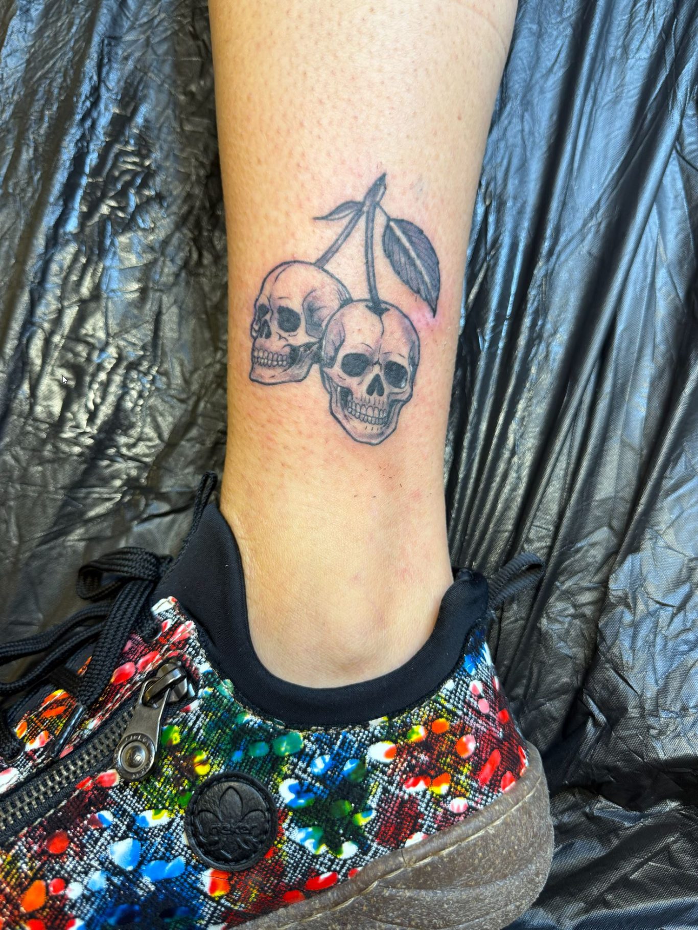 Paradize INK Tattoo Mömlingen Tätowierung von zwei Schädeln mit einer Pflanze auf einem Knöchel. Glitzernde Schuhe im Hintergrund.