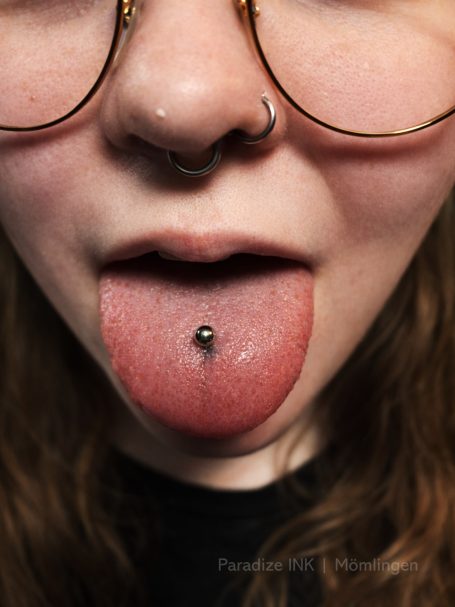 Nahaufnahme eines Gesichts mit Brille, Zunge herausgestreckt und Piercing im Lippenbereich.