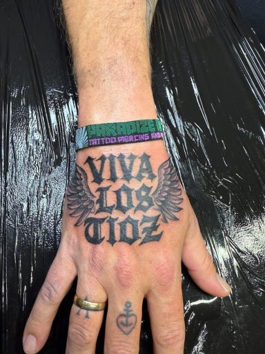 Tattoo auf einer Hand mit dem Schriftzug "Viva Los Tioz" und Flügelmotiv.