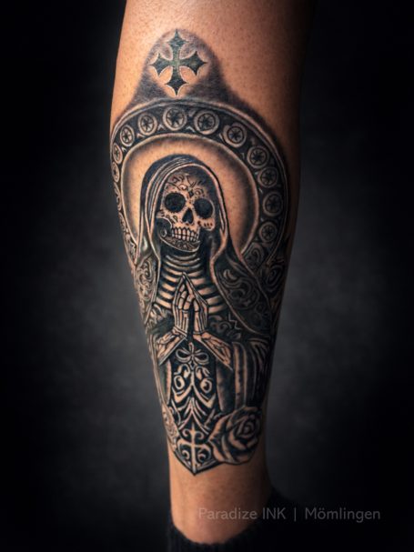 Schwarzes Tattoo eines Totenkopfes in der Pose einer betenden Figur mit Ornamenten.