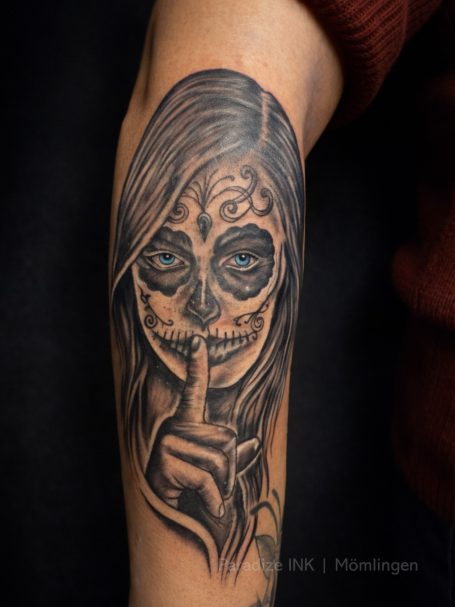 Tattoo eines Gesichts mit auffälligen Augen und einem Finger vor dem Mund zum Schweigen.