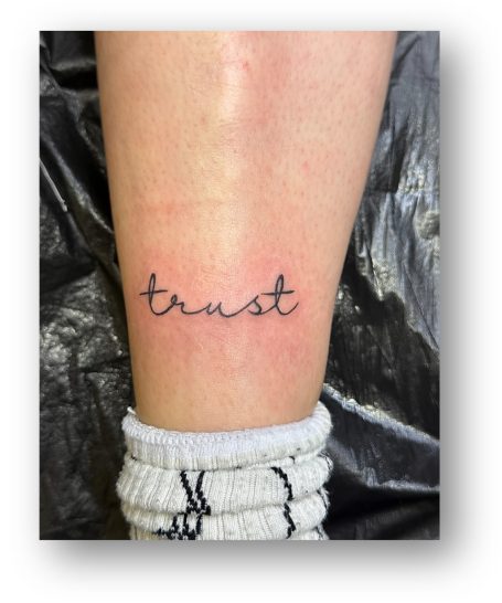 Kleine Tätowierung am Bein mit dem Wort "trust" in geschwungener Schrift.