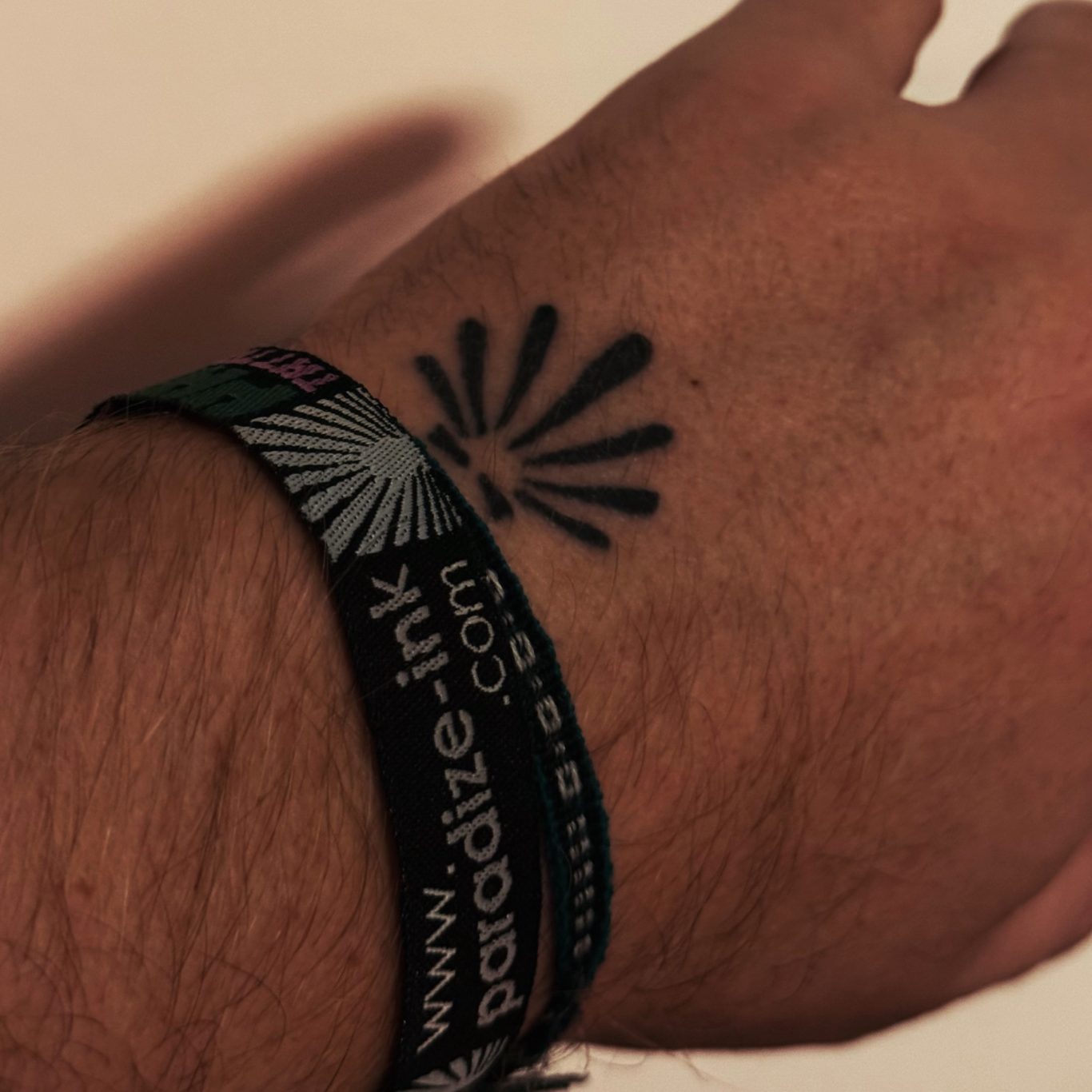 Paradize INK Tattoo Mömlingen Ein Handgelenk mit einem schwarzen Tattoo und einem bunten Festivalarmband.
