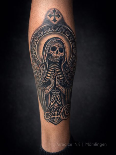 Tattoo eines Skelets in einer religiösen Pose, umgeben von dekorativen Mustern.