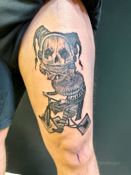 Tattoo auf dem Oberschenkel: Totenkopf mit Jester-Maske und ornamentalem Design.
