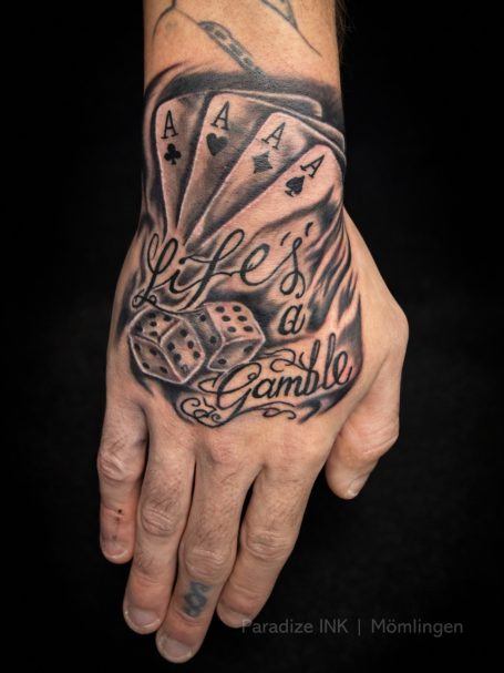 Tattoo auf einer Hand mit Würfeln und Spielkarten, der Schriftzug „Life's a Gamble“.