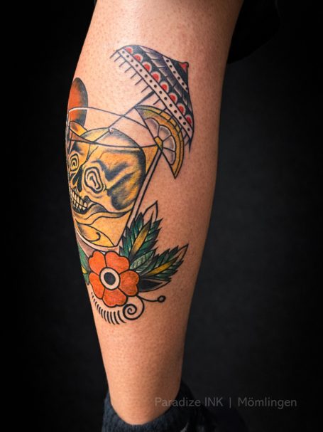 Buntes Tattoo eines Cocktails mit Schirm und Blumenmotiven auf einem Unterschenkel.